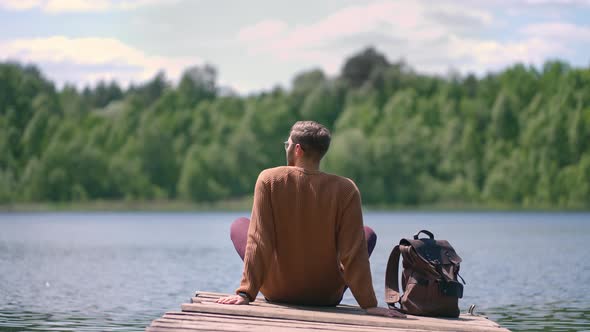 One Man with a Backpack Rests Sitting on a Pier on the Shore of a Lake in the Woods