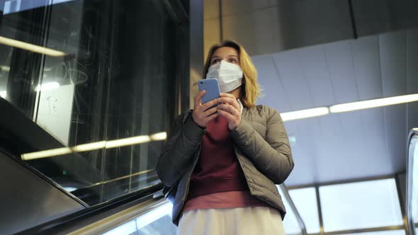 Young Woman Wearing Medical Mask Stands on Moving Escalator in Subway and Using Smartphone