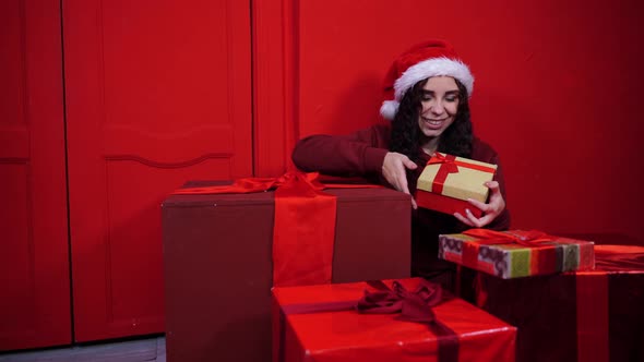 Young Woman in Santa Claus Hat Sitting in Gifts Near Red Wall