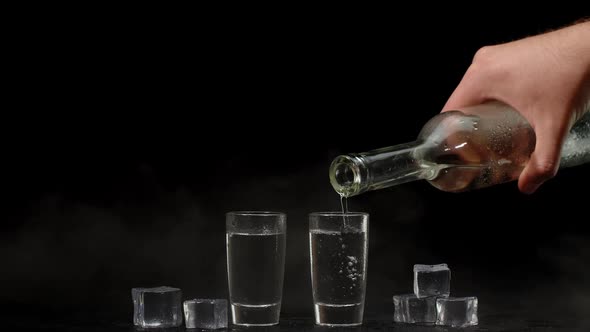 Bartender Pouring Up Vodka From Bottle Into Two Shots Glasses with Ice Cubes on Black Background