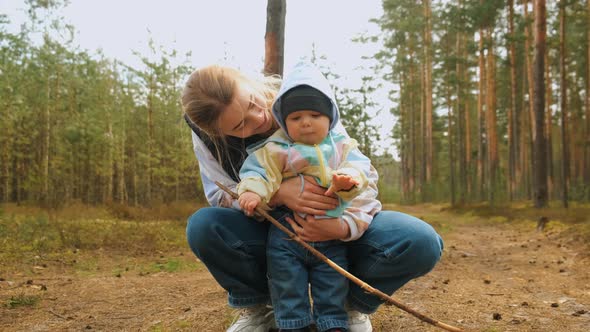 Little Baby Boy Holding a Big Stick Walks Around the Forest Road with the Help of His Mum