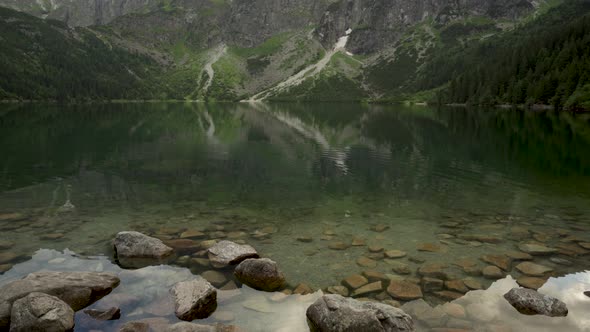 Tilt Up Shot Of Rocks In A Clear Lake Against Mountains In Morskie Oko