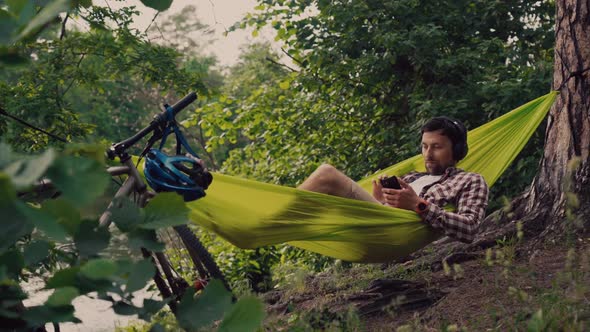 Man on Bicycle Trip at Camping By Lake is Relaxing in Green Hammock While Listening to Music