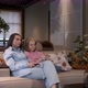 Happy young woman is sitting on cozy sofa with adorable little cute daughter. Watching TV - VideoHive Item for Sale
