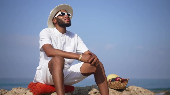 Handsome Confident Male African American Tourist Sitting on Cliff with Clear Blue Summer Sky and