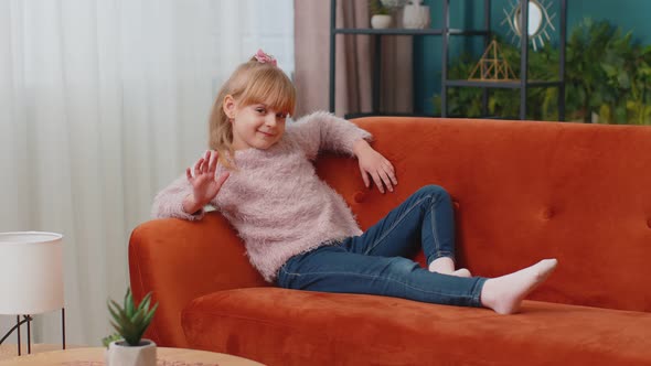 Child Girl Lying on Sofa at Home Looking at Camera Smiling Waving Hands Gesturing Hello or Goodbye