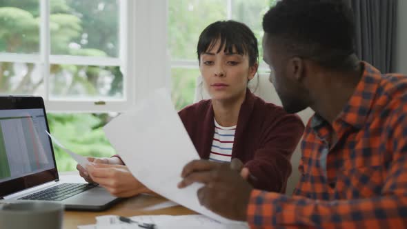 Diverse couple sitting at table talking and working with laptop