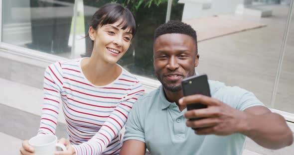 Happy diverse couple wearing blouse and shirt and taking selfie with smartphone in garden