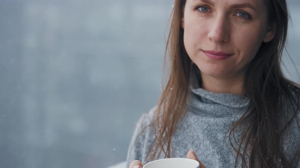 Caucasian Woman Stays on Balcony During Snowfall with Cup of Hot Coffee or Tea