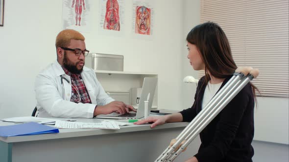 Young Woman with Crutches Visiting Afroamerican Male Doctor