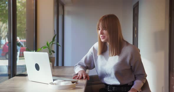 Young Sociable Blonde Cheerfully Speaks By Video Chat on a Laptop While Sitting in a Cozy Cafe