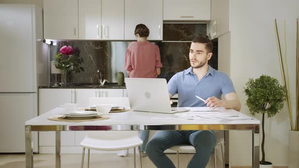 An Adult Man is Sitting and Working at Home and His Wife is Giving Him a Cup of Drink