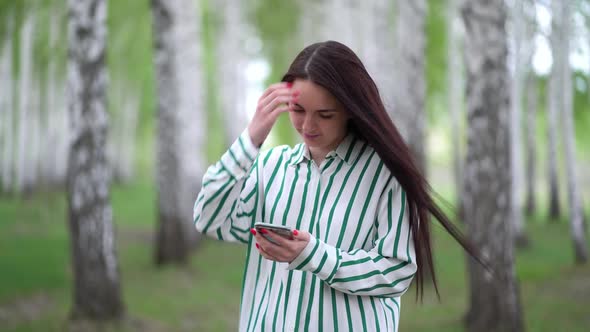 Beautiful Girl with a Smartphone in Her Hands Walks Along a Birch Grove in Spring