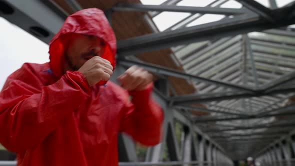 a Man in a Red Jacket with a Hood is Boxing on an Iron Bridge