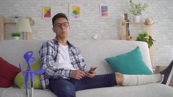 Sad Asian Young Man with a Broken Leg in Bandages Lying on the Couch Uses Smartphone and Looking at