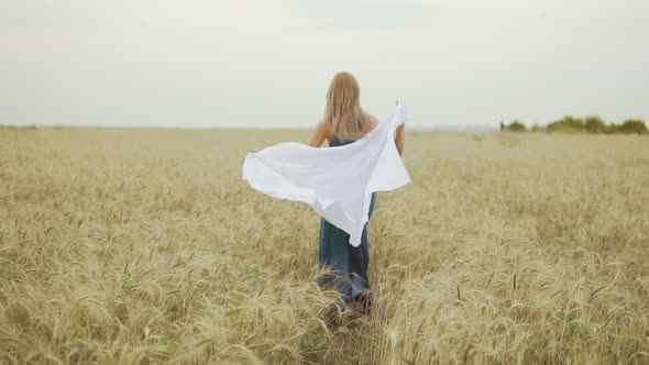 Back View of Attractive Young Woman in a Long Blue Dress Running Through Golden Wheat Field Holding