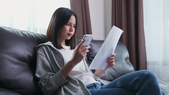 Asian woman holding the bill and smartphone in the living room and stressed by excessive expenses.