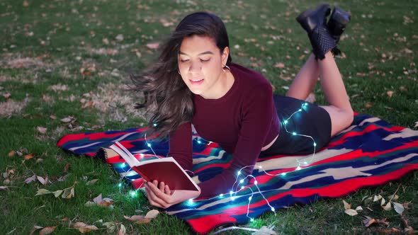 An attractive young woman reading a story book or novel in a park at twilight with lights glowing ar