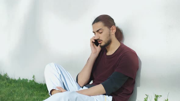 Portrait of Young Attractive Arab Man Sitting on the Grass and Talking on the Phone