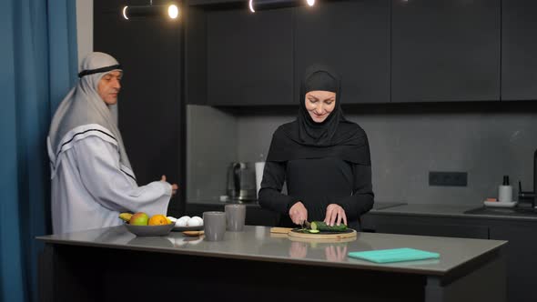 Charming Slim Middle Eastern Woman Slicing Cucumber for Healthful Salad As Smiling Man Entering