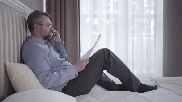 Senior Businessman Sitting on Bed in Hotel Room and Talking on the Phone. Confident Adult Caucasian