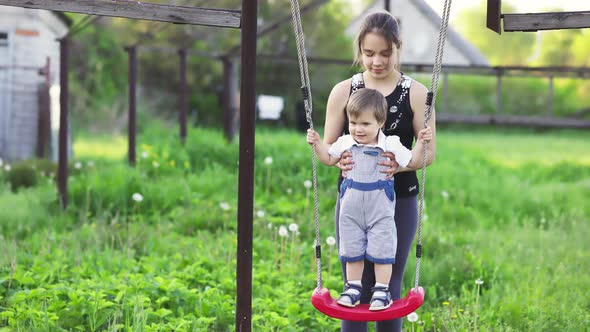 Cute Brother and Older Sister Ride on a Bright Swing on Warm Spring Day Against the Backdrop of a