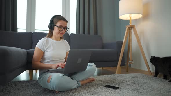 Casually Dressed Woman with Headphones is Sitting on Carpet with Laptop and Working in Cozy Room