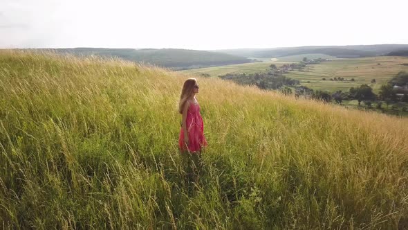 Yong Woman with Long Hair in Red Dress Walking in Summer Field with Tall Green Grass on Rural Hill