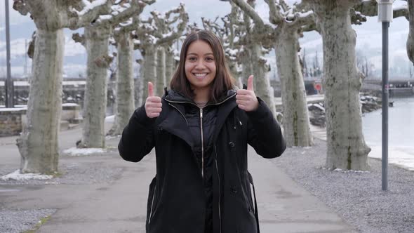 Young model woman shows thumbs up while standing in wintry city landscape between trees.