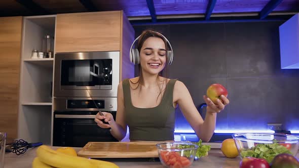 Young Woman which Eating Apple while Listening Music in Headphones in the Kitchen