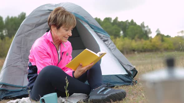 Senior Woman Reading a Book While Camping Outdoor with Tent in the Background