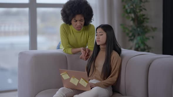 Sad Young Asian Woman Sitting on Couch with Laptop As African American Friend Calming Down