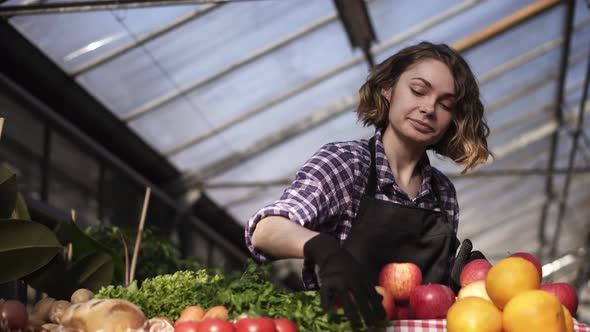 Low Angle View of a Beautiful Smiling Woman Farmer in Black Gloves Arranging Organic Food in Farm