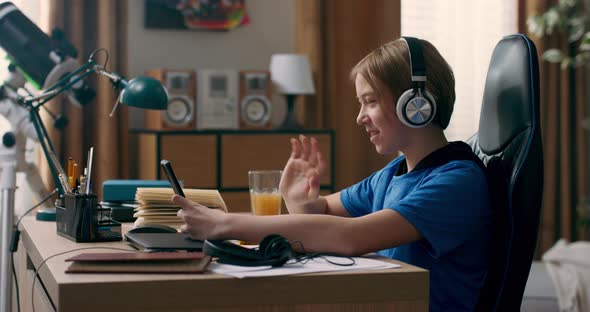 Young Boy Sits in Room in Morning Drinking Orange Juice Holding Phone in Hands Watching Videos alt