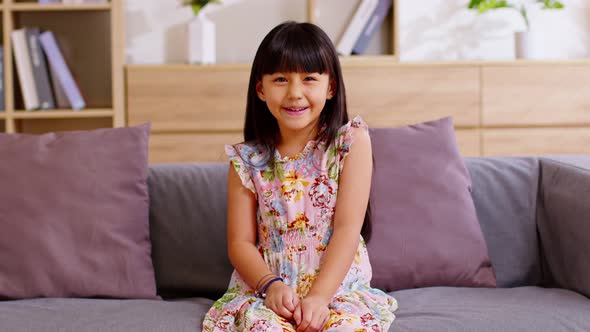 Cute Asian girl smiling at the camera while sitting on couch in the living room. She is pretty, cute