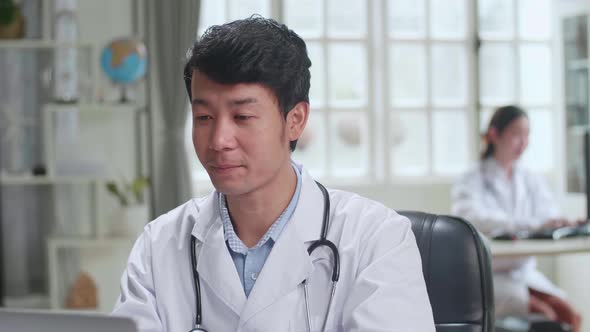 Asian Male Doctor Communicates With Patient By Video Link on Laptop Computer In Workplace