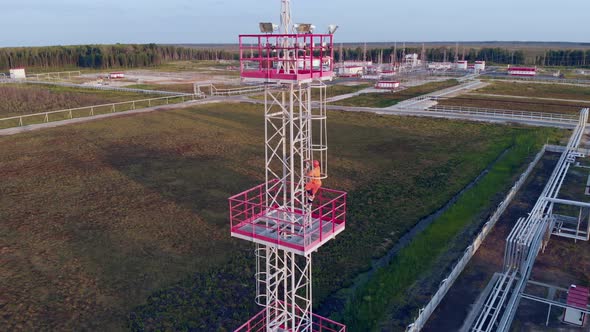 A Worker in a Helmet and Overalls Climbs an Iron Ladder to a Metal Tower of Illumination at a Power