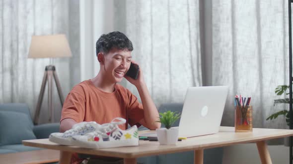 Asian Boy Footwear Designer Talking On Smartphone While Working On A Laptop At Home