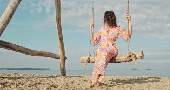 Young Girl in Dress Sways on Swing on Seashore Ocean