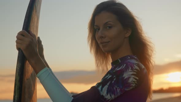 Young Attractive Female Surfer Smiling and Posing with Her Surfboard at Sunset on the Beach