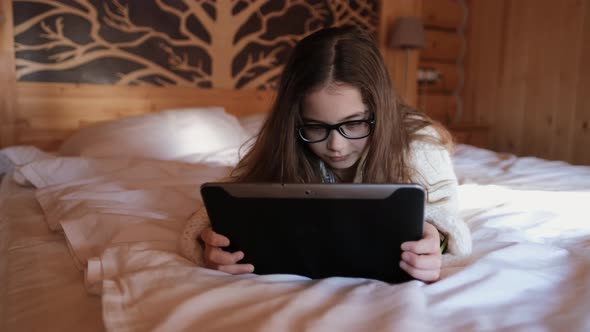 Cute Teenage Girl Browses the Internet on a Tablet Lying on a Bed in a Country Cozy House