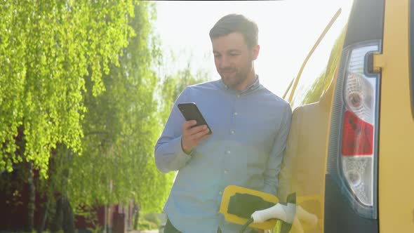 Young Man Chat on Mobile Phone While Electric Car is Charging on Outdoor Station