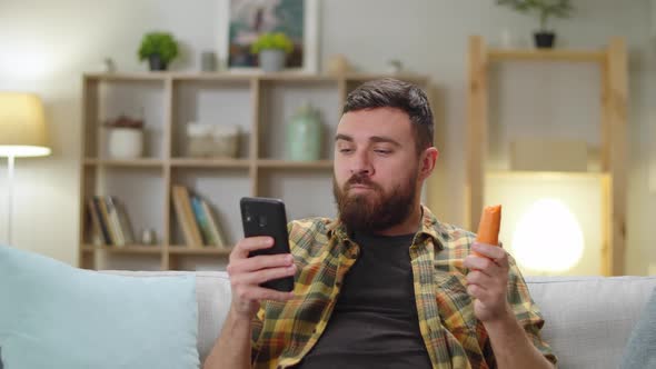 Young Handsome Caucasian Male Eating Carrot at Home in Living Room Sitting on Sofa with Phone
