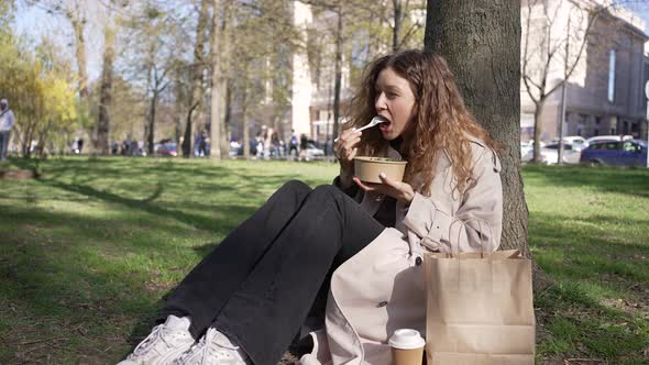 Young Woman Enjoys Lunch Leaning on Tree in City Park