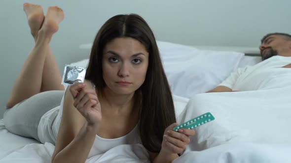 Puzzled Woman Holding Condom and Oral Pills, Contraception Choice, Health Care