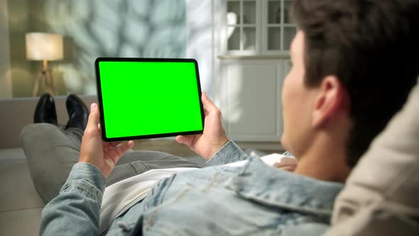 Over The Shoulder View Young Man at Home Lying on a Sofa Using With Green Mock-up Screen Tablet