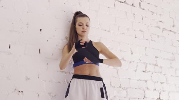 A Girl Boxer Against a White Brick Wall Wraps a Sports Bandage for Boxing on Her Hands