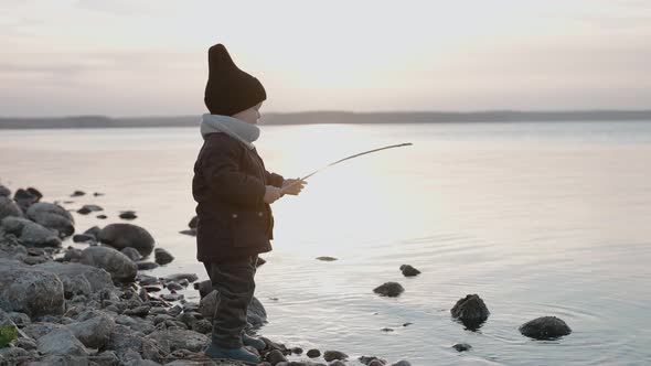 Baby Boy Playing with Stick and Water on the Stone Shore on an Autumn Evening