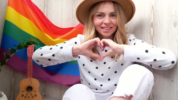 Young Blonde Millennial Hippie Woman Showing Heart Sign with Hands with Rainbow LGBTQ Flag at Home