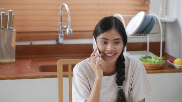 Young Asian woman talking on the phone while having breakfast in the kitchen.